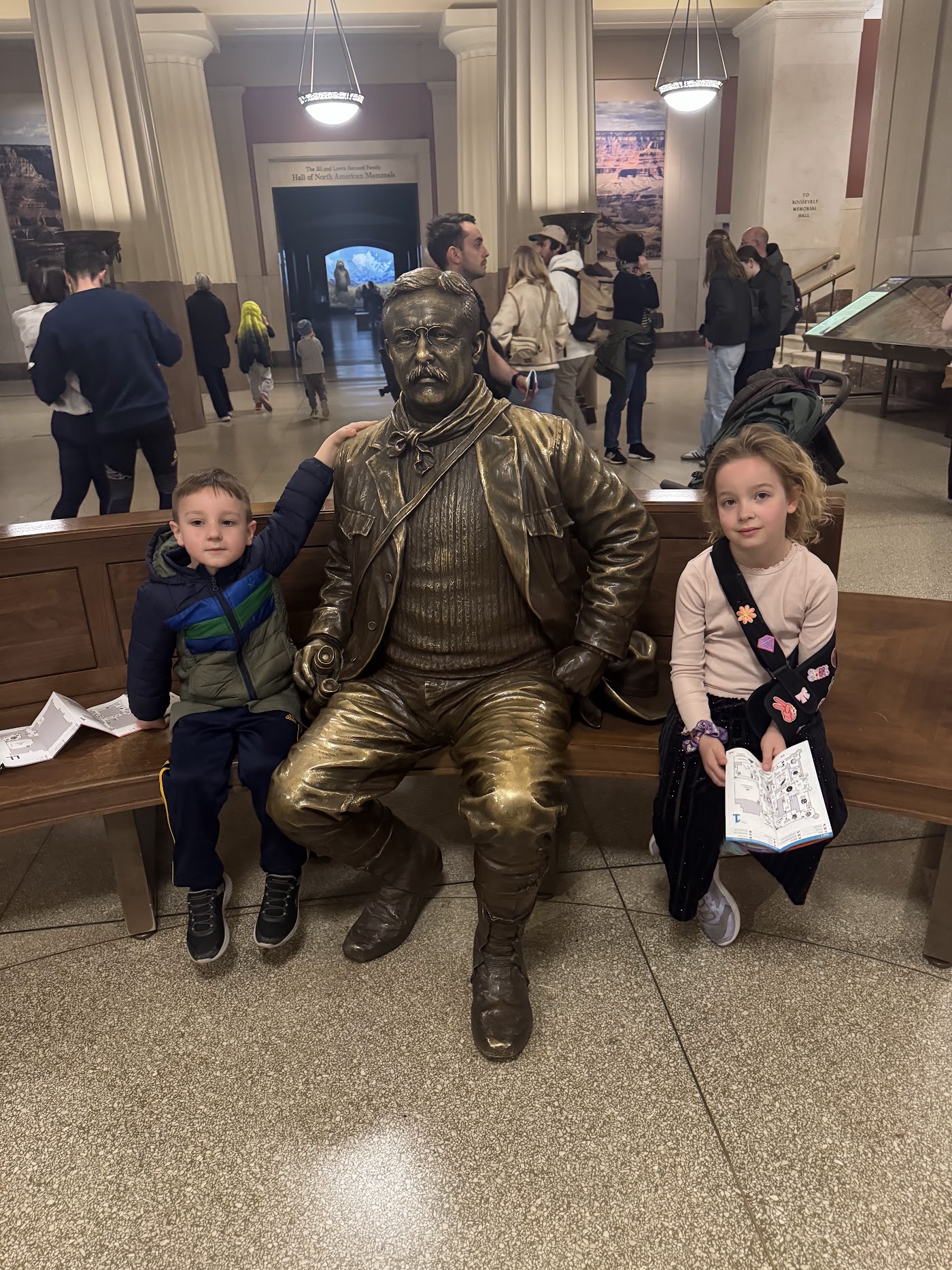 Two children on a bench either side of a seated bronze statue in a museum hall.