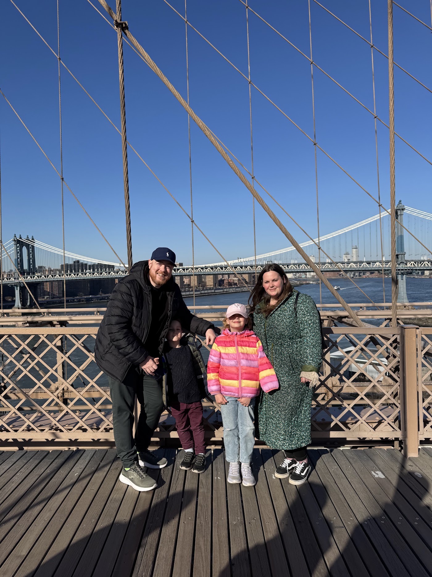 A family of four, two adults and two children, on the wooden walkway of the Brooklyn Bridge under its cables.