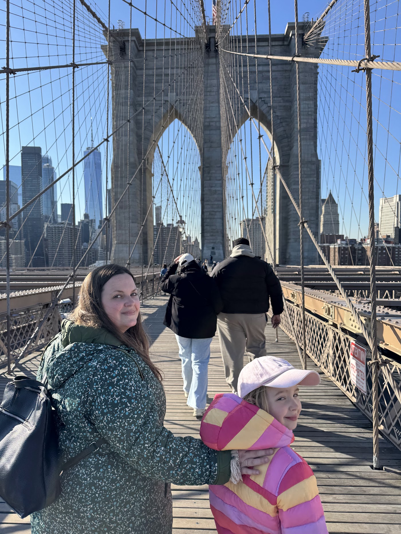 A woman and two children walking on the Brooklyn Bridge boardwalk with the stone tower and cables ahead.