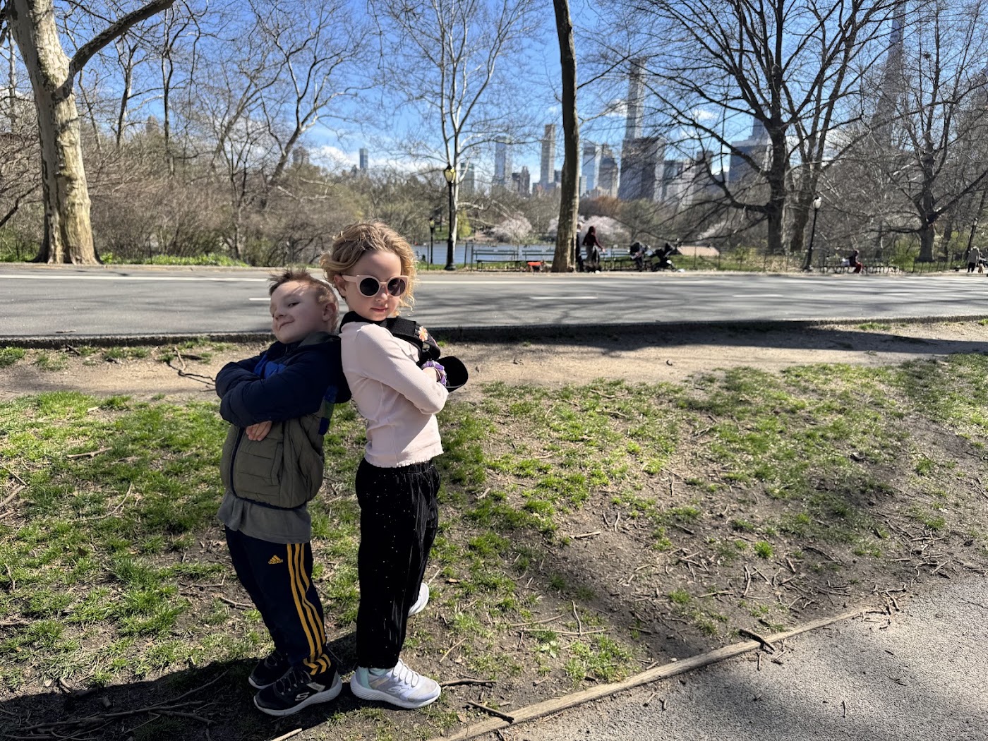 Two children hugging on the grass, bare trees and tall buildings behind them.