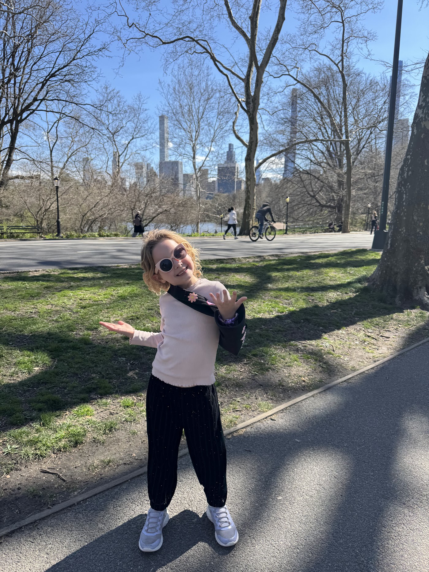 A girl in sunglasses on a park path, skyscrapers in the distance.