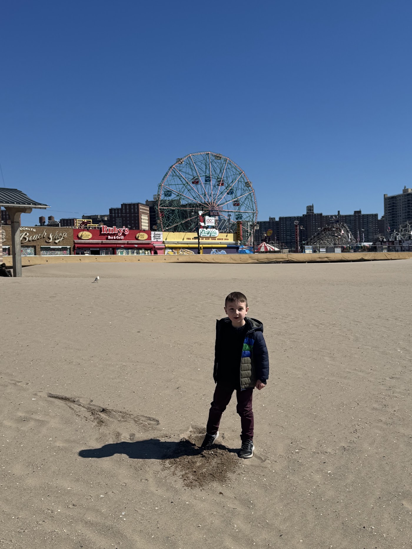 A child on a wide empty beach with the Wonder Wheel and amusement park in the background.