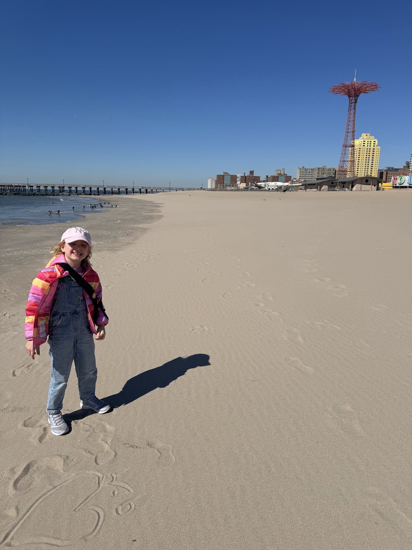 A child in a pink coat walking on the beach, the red Parachute Jump tower behind.