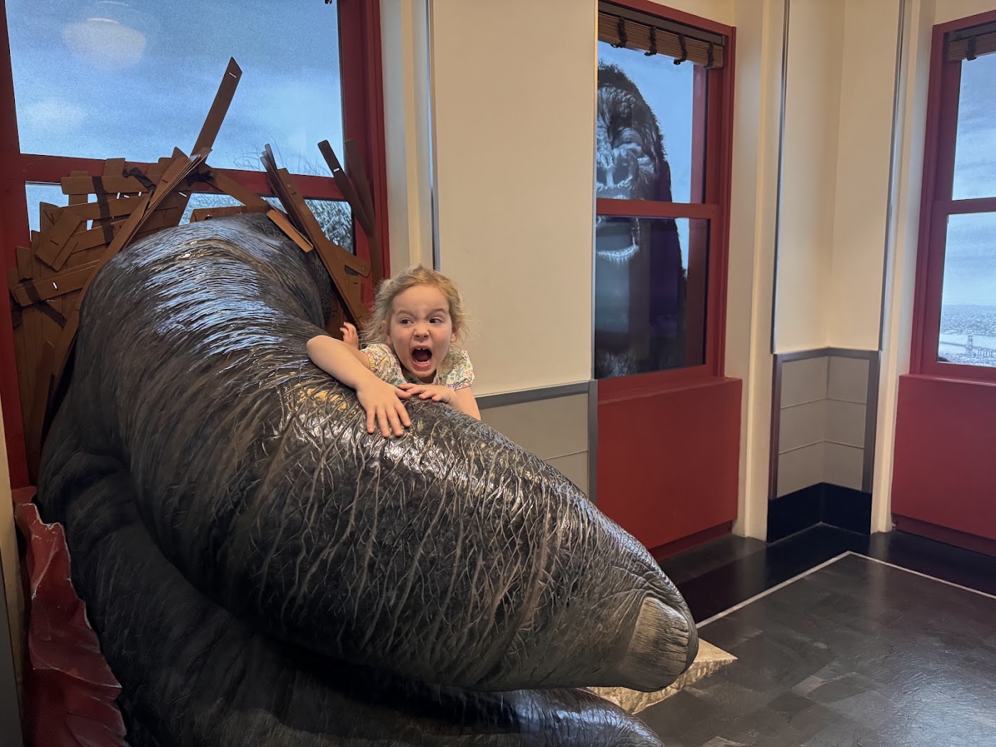 A child sitting on huge model ape fingers that reach through a window frame.