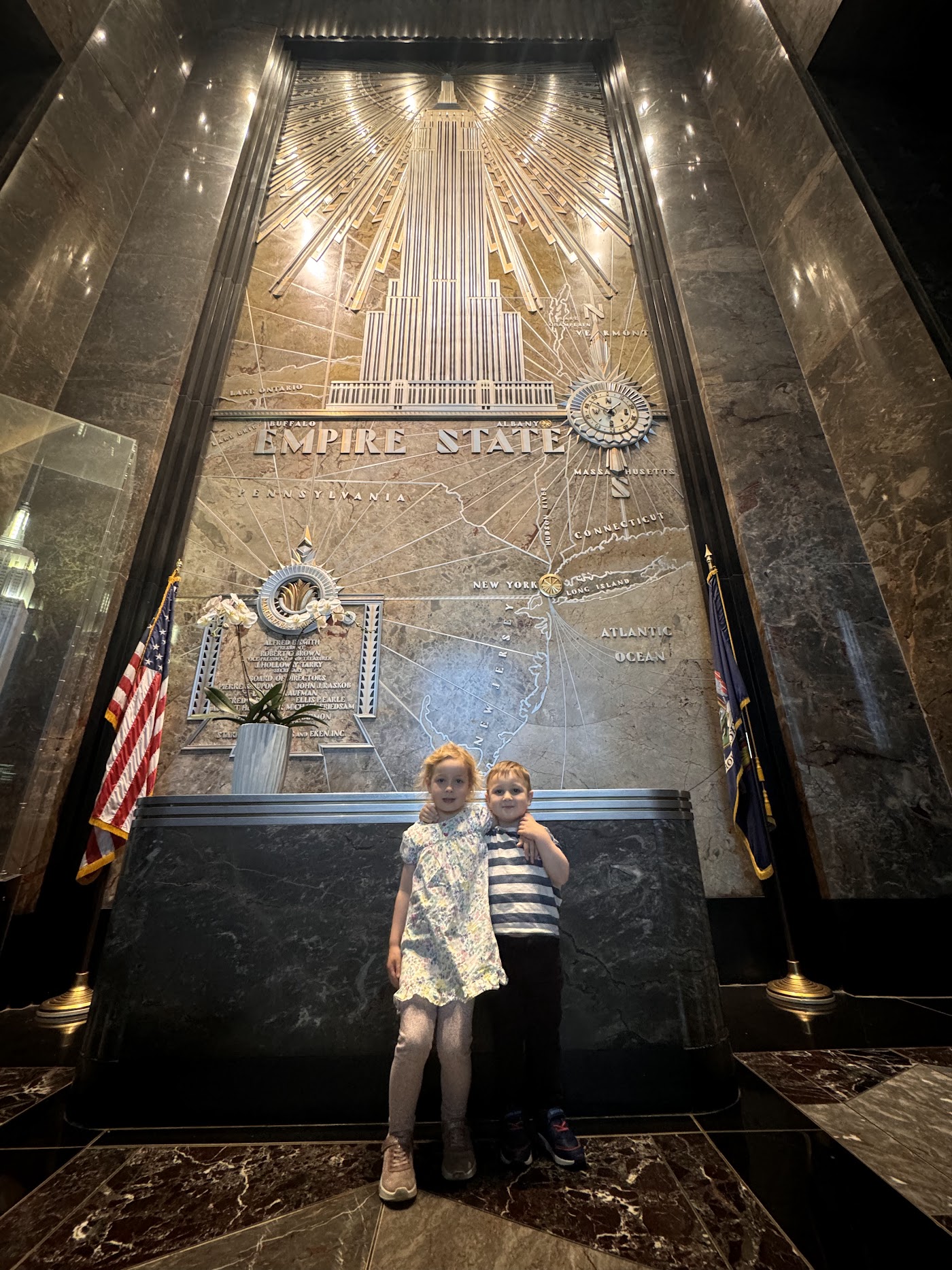 Two children in a marble lobby below a gold mural of the Empire State Building.