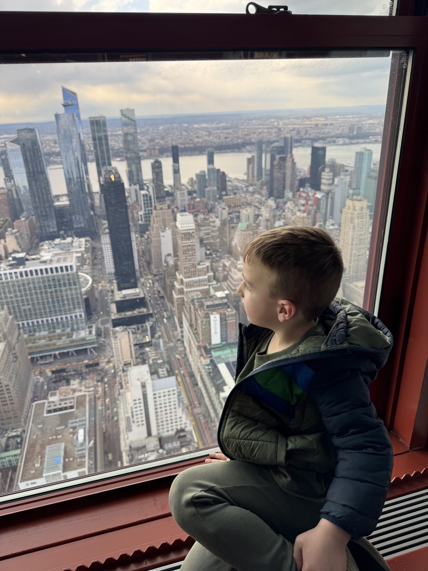 A boy looking out of a tall window over Midtown rooftops and the skyline.