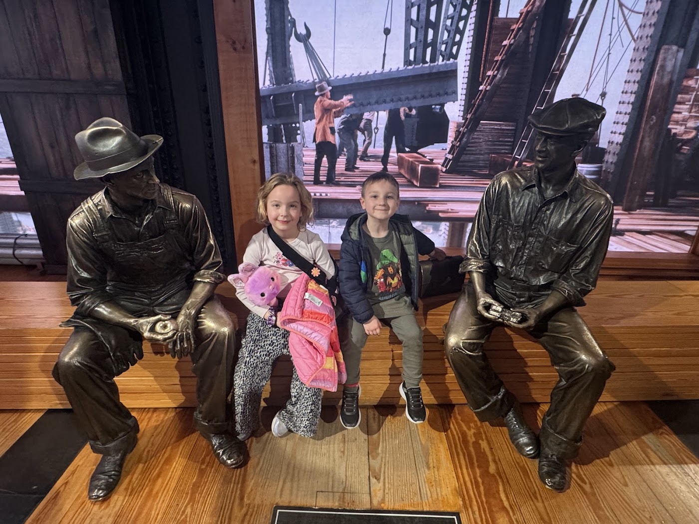 Two children on a bench between two bronze statues of construction workers.