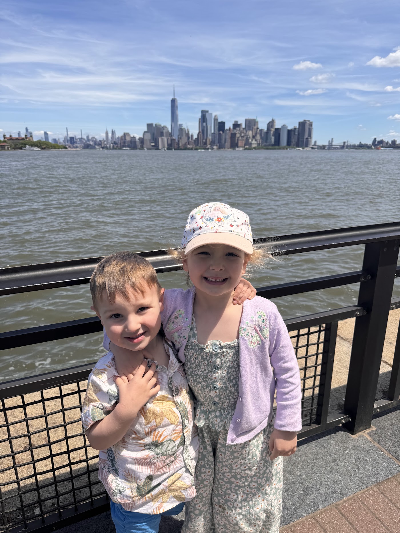 Two children at a railing on Liberty Island, the skyline behind them under a blue sky.