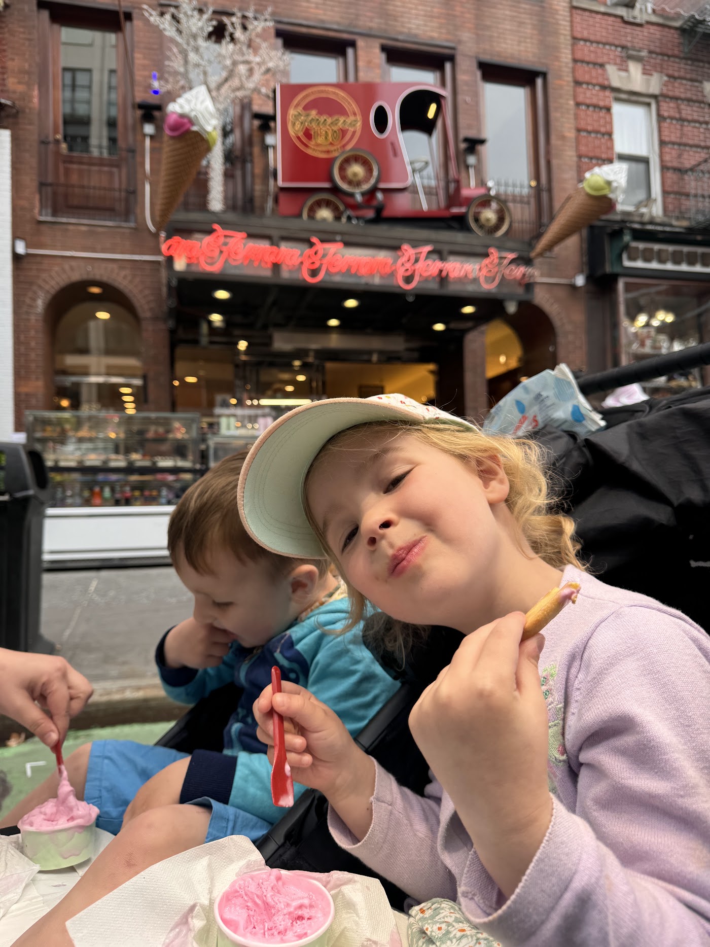 Two children at a cafe table outside with ice creams, a restaurant behind them.