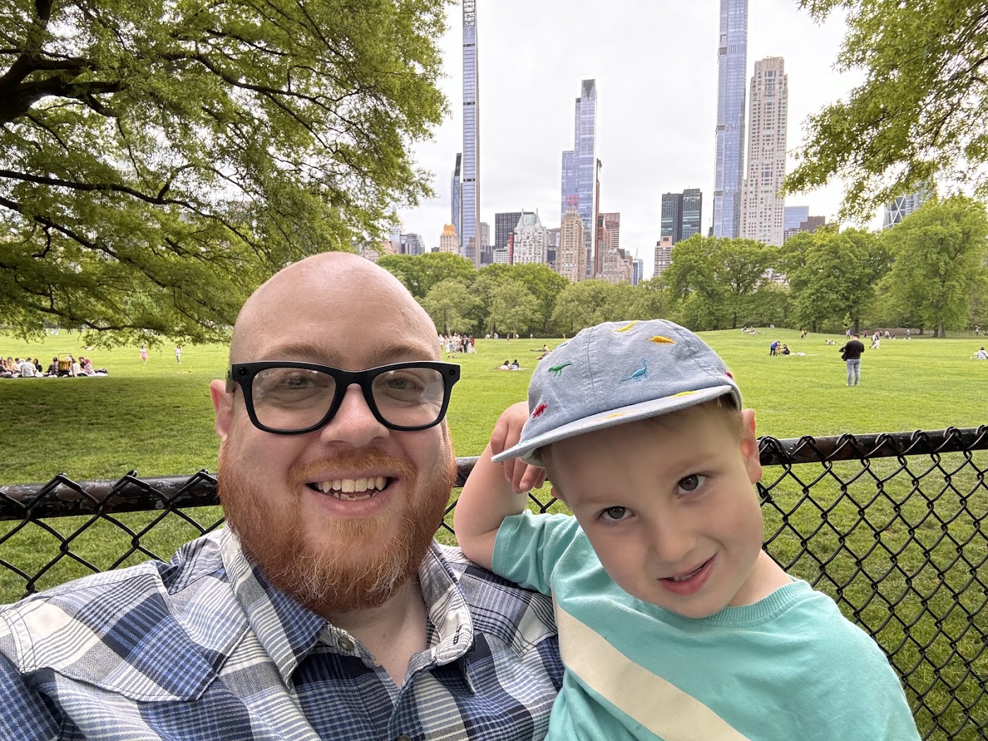 A dad and a small boy in a cap on the grass, with tall thin skyscrapers in the background.