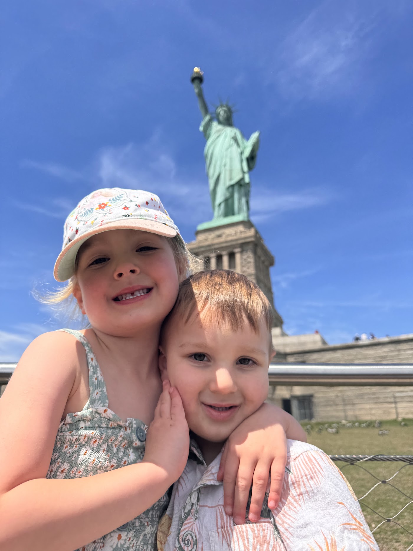 Two children in sun hats grinning, with the Statue of Liberty rising behind them.