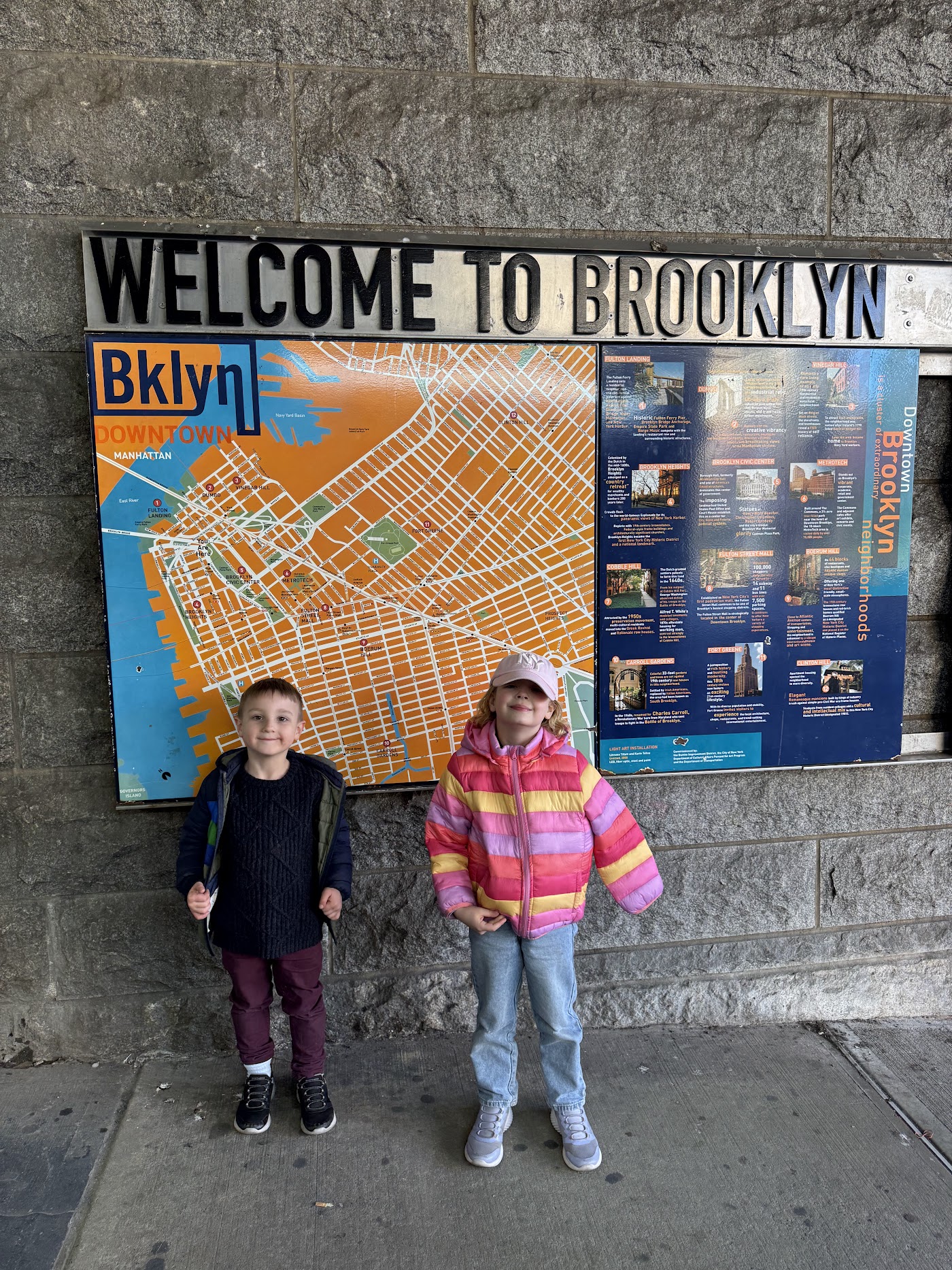 Two children in front of an orange and blue Welcome to Brooklyn map sign.